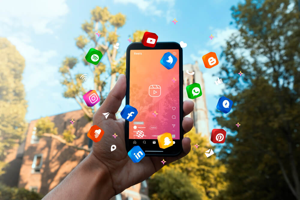 Hand holding smartphone outdoors displaying a reels-style screen while colorful social media app icons float around against sunny trees.