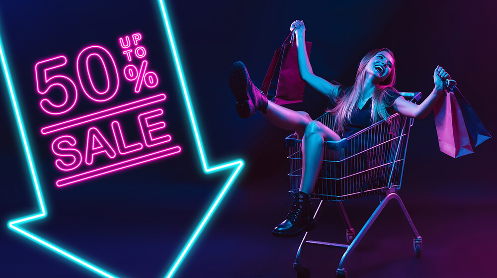 Young woman joyfully sits in a shopping cart holding colorful bags beside neon 'Up to 50% Sale' arrow, vibrant magenta and cyan lighting.
