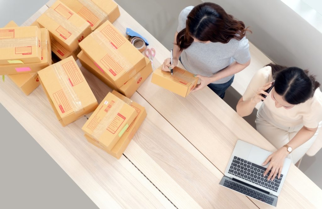 Two women preparing ecommerce packages with a laptop, tape, and multiple cardboard boxes on a wooden table 